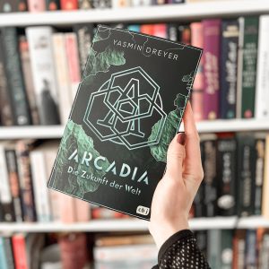Das Buch Arcadia -Die Zukunft der Welt wird in der Hand gehalten. Im Hintergrund ist verschwommen ein Bücherregal.